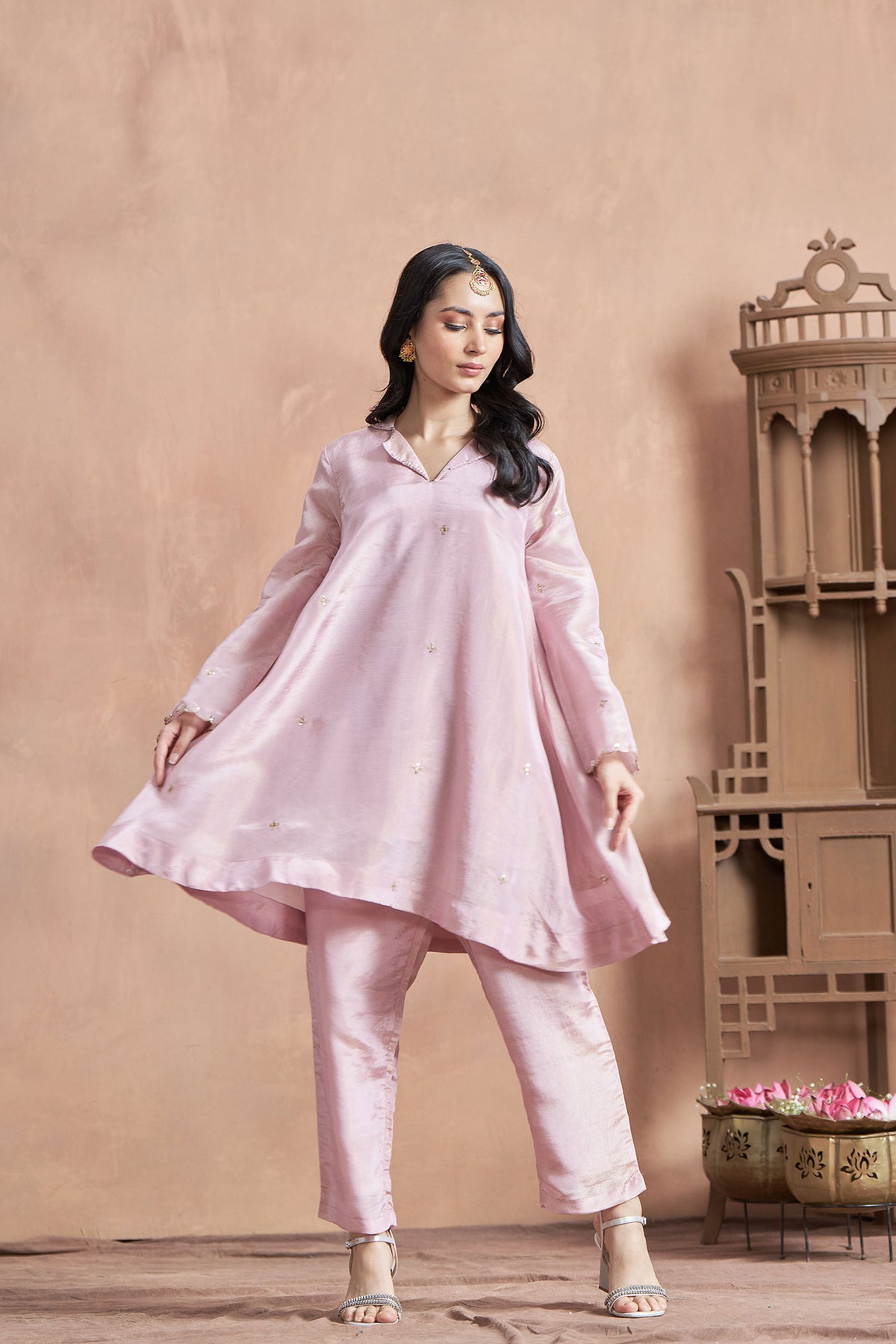 A woman wearing a loose-fitted pink shimmer tissue co-ordinate set, consisting of a top and pants, with details on the sleeves and neckline.