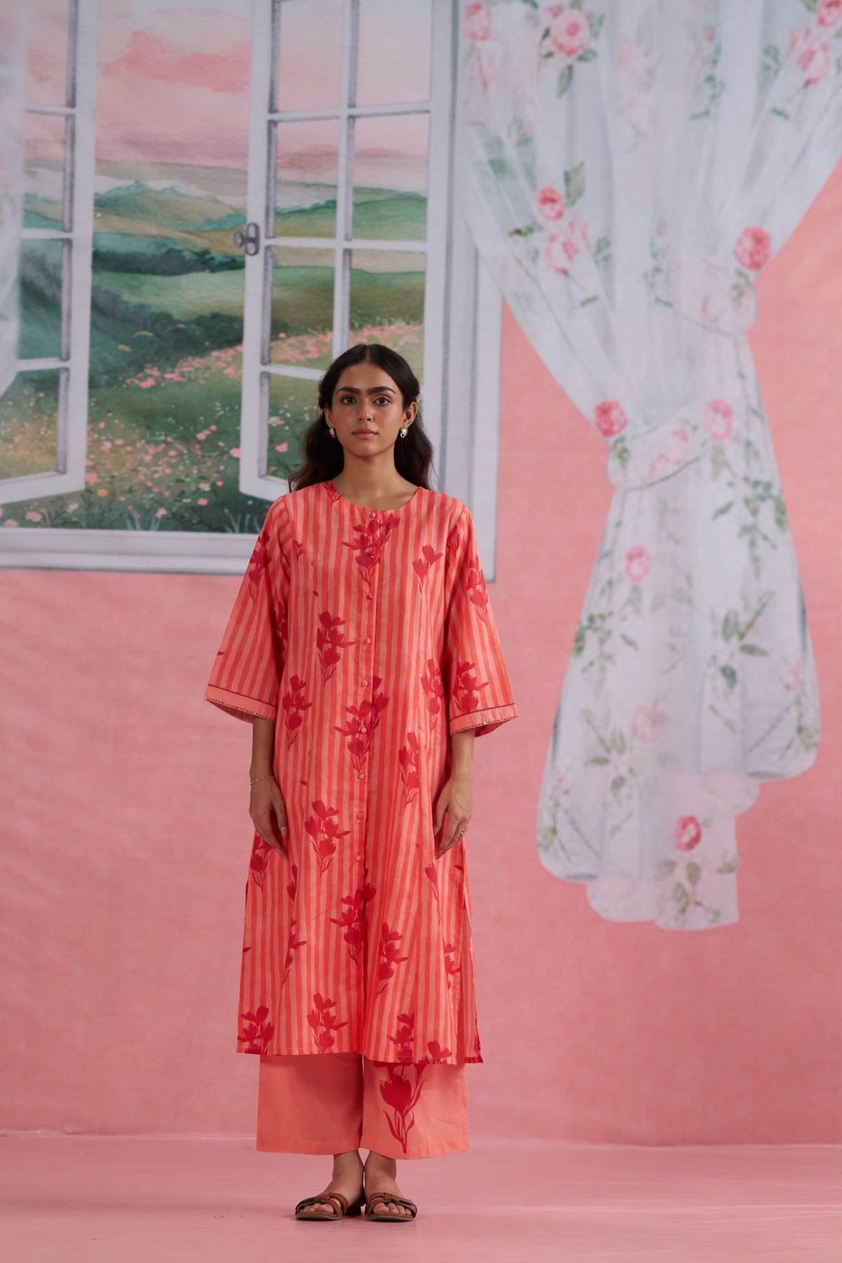 Woman wearing a red floral outfit standing in front of a pink wall with a window and curtain.