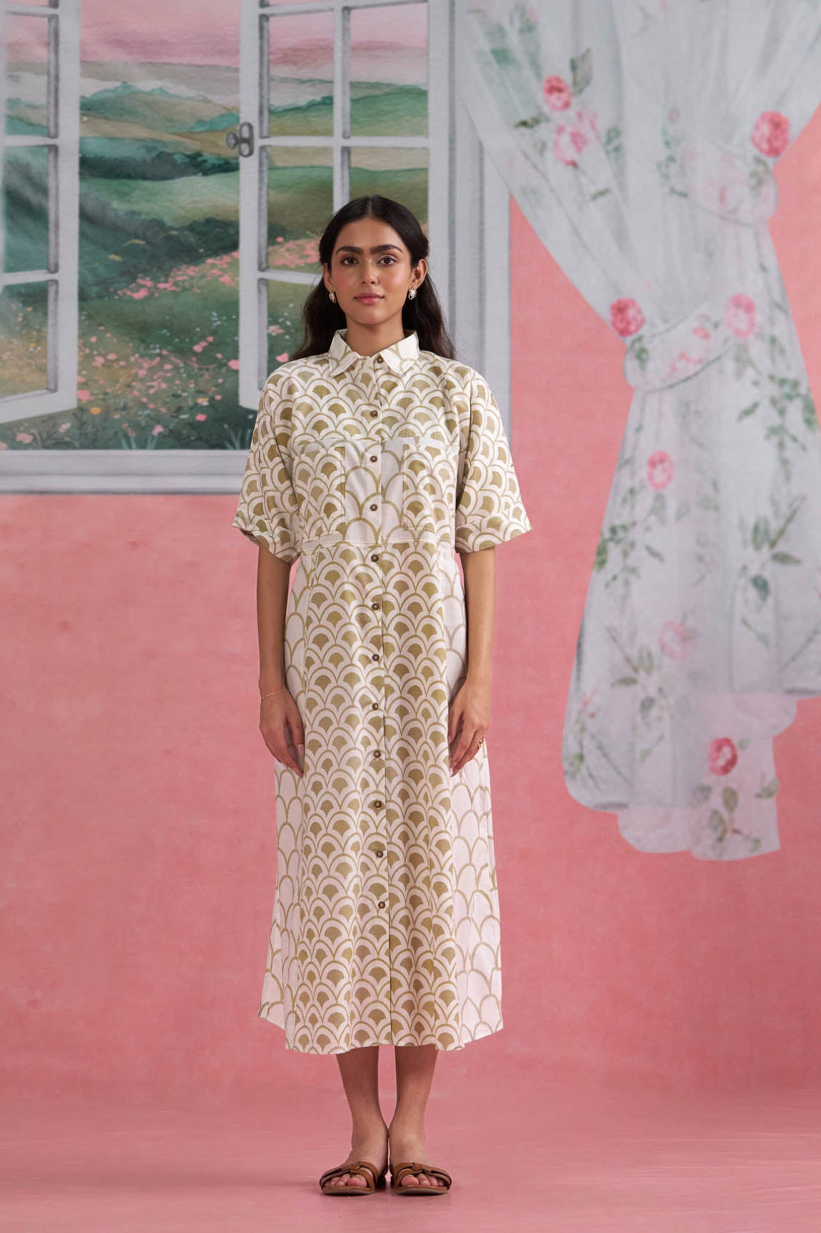 Woman in a floral dress standing in front of a painted window scene on a pink wall.