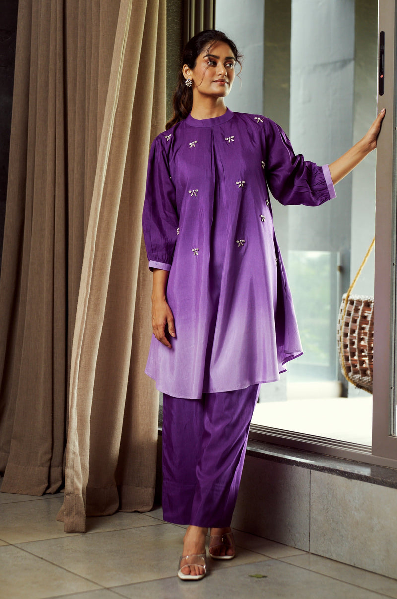 Woman in a purple outfit standing by a window with curtains.